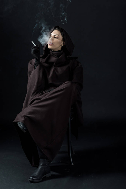 Nachdenkliche Frau im Todeskostüm sitzt auf Stuhl und raucht Zigarette auf Schwarz - Foto, Bild