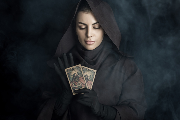 KYIV, UKRAINE - APRIL 18, 2019: attractive woman in death costume holding tarot cards on black - Zdjęcie, obraz