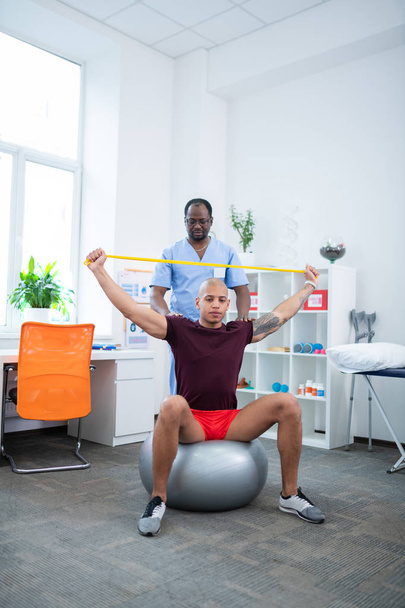 Mann sitzt auf Fitnessball und streckt Arme zum Therapeuten - Foto, Bild