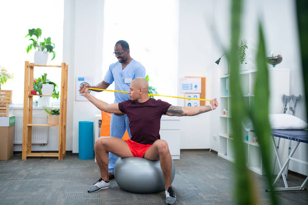 Bodybuilder sitzt auf Fitnessball und arbeitet mit Therapeut - Foto, Bild