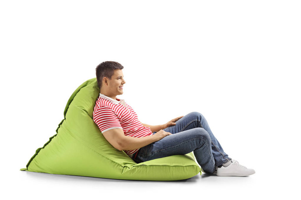 Full length profile shot of a young man sitting on a green bean bag and smiling isolated on white background - Foto, Bild