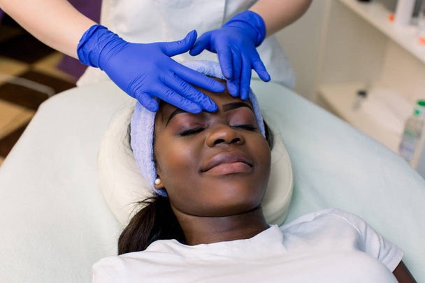 Relaxed Young African Woman Receiving Forehead Massage In Spa. Woman massagist in blue rubber gloves performing facial massage. Spa, massage concept - 写真・画像