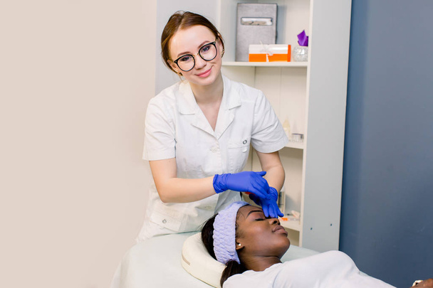people, beauty, spa, lifestyle and relaxation concept - young smiling woman beautician in medical gloves doing face massage to beautiful young African woman at spa - Photo, image