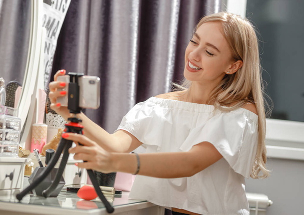 Jolie fille blogueuse pose le téléphone sur le stand sur la coiffeuse pour le tournage vidéo pour son blog
 - Photo, image