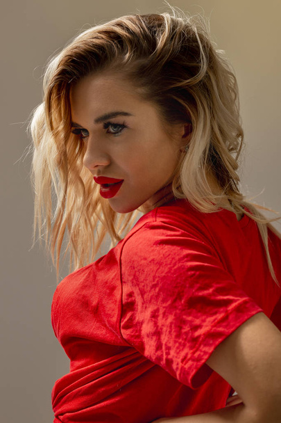 Blond slim girl with red lipstick dressed in a red T-shirt is posing against a wall in the studio - Foto, Imagen