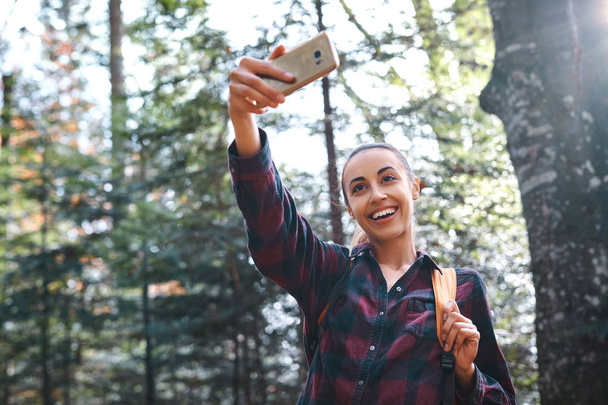 Beautiful happy stylish traveling woman taking photo on the camera phone with forest background. - Photo, Image