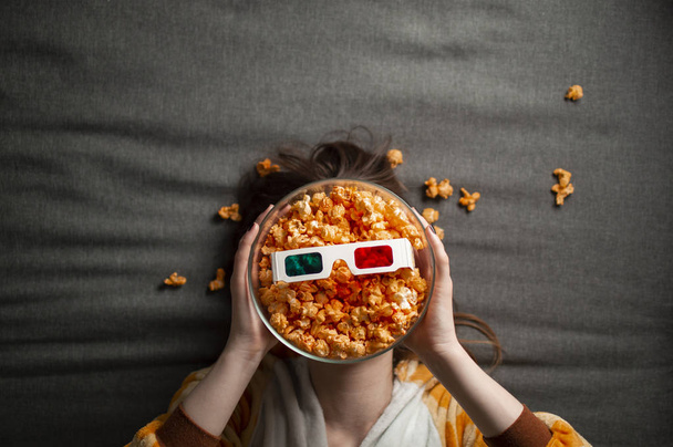 молодая милая девушка лежит на сером фоне в 3D очки ест попкорн и смотрит фильм
 - Фото, изображение