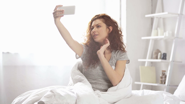 piękna dziewczyna pokryta kocem biorąc selfie na smartfonie i uśmiechając się w łóżku  - Materiał filmowy, wideo