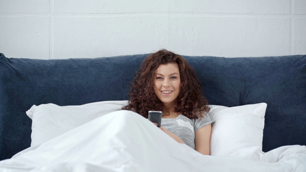 mooi meisje liggend in bed met afstandsbediening, glimlachend en tv kijken thuis - Video
