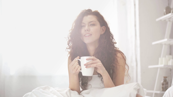 beautiful girl sitting on bed, drinking coffee, looking at camera and smiling in morning - Filmati, video