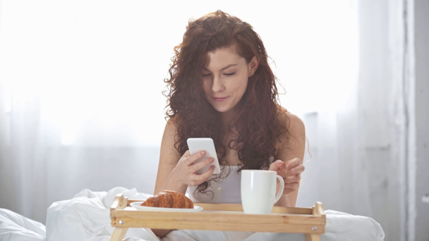 hermosa chica usando teléfono inteligente, beber café, sonriendo y mirando a la cámara en la cama
 - Metraje, vídeo
