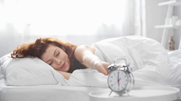mooi meisje liggend in bed, kijkend naar wekker, glimlachend en stretching in de ochtend - Video