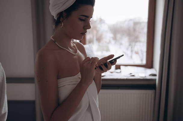 schoonheid vrouw in een badjas en met een handdoek op haar hoofd met behulp van smartphone - Foto, afbeelding