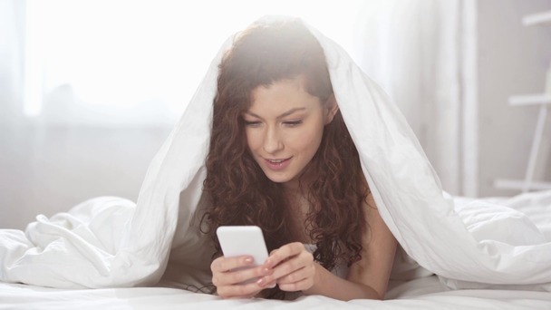 beautiful girl covered with blanket typing on smartphone and smiling in bed  - Imágenes, Vídeo