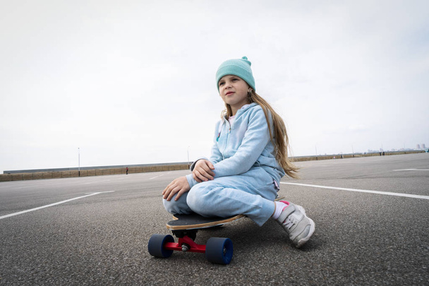 A girl in a blue hat and sweater sits on a large longboard along a large asphalt ground. - Zdjęcie, obraz