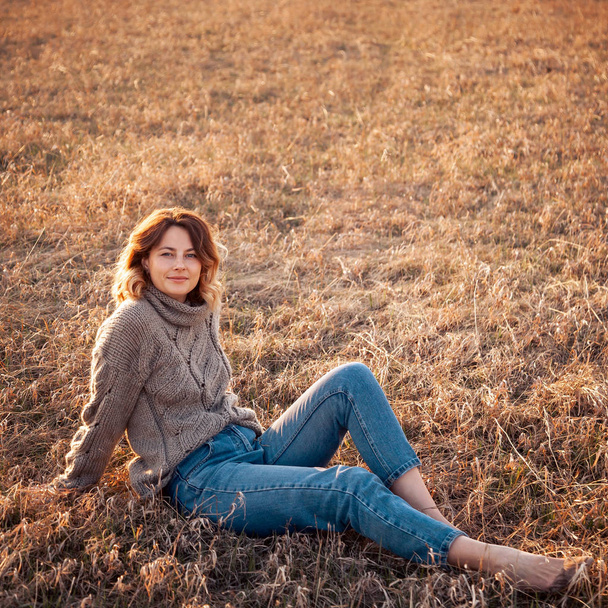 Portrait of a beautiful young model in warm clothes enjoy day, on background field in  sunny autumn day . Autumn warm photo. Woman smiling and look away, joyful cheerful mood. - 写真・画像