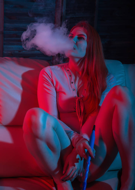 Sexy redhead woman vaping electronic hookah in red blue tones - Photo, Image