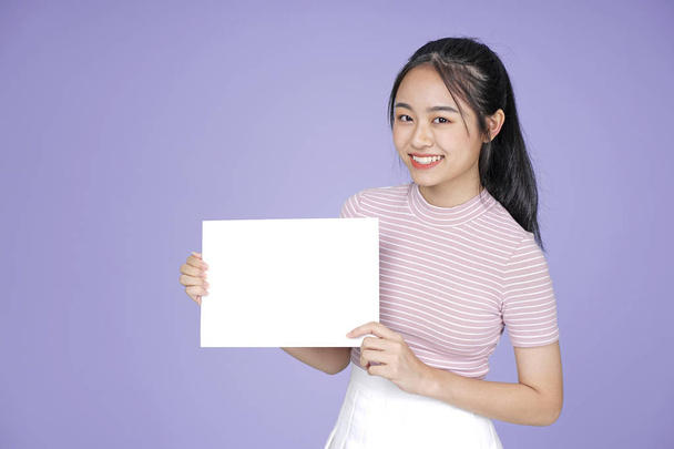 Asian young teenage beautiful cheerful girl with blank message white board in pink shirt, on violet background - Φωτογραφία, εικόνα