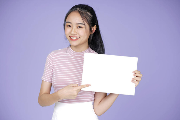 Asian young teenage beautiful cheerful girl with blank message white board in pink shirt, on violet background - Fotoğraf, Görsel