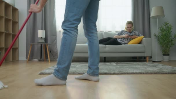 Tired man mopping parquet floor and his girlfriend play on smartphone on sofa - Materiał filmowy, wideo