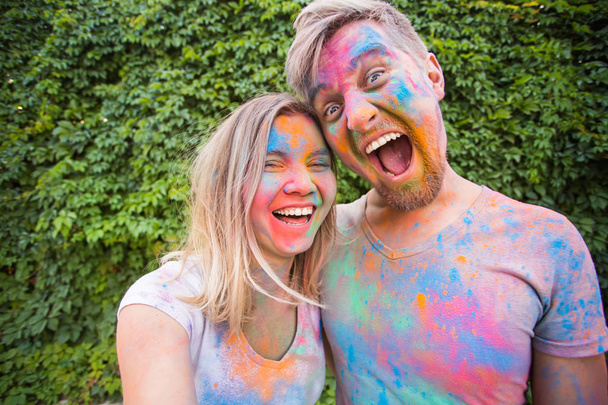 Holiday, holi and people concept - Woman and man with painted faces taking selfie over bush backgrund - Photo, Image