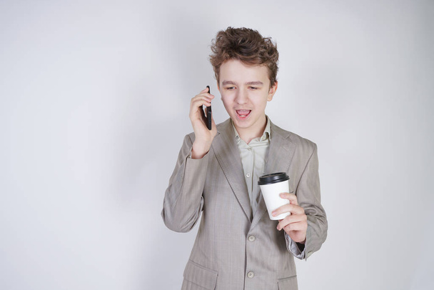 caucasian teenager in a gray business suit stands with a mobile phone and a paper Cup of coffee on a white background in the Studio - Photo, Image