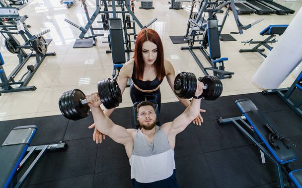 Top view of a strong man lifting dumbbells and a pretty woman helps him in gym. Active couple exercising with dumbbells on the background of a modern sports club. - Фото, изображение