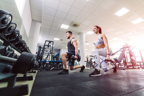 Healthy couple in sport clothes lifting dumbbells in the gym. Attractive woman and handsome man doing workout with dumbbells standing in special pose in the sports club. - Fotoğraf, Görsel