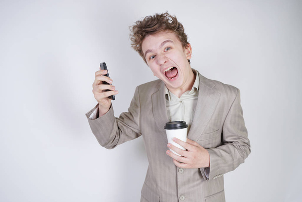 aggressive caucasian teenager screams into the smart phone and holds a paper cup of coffee in his hand. student on edge on a white background in Studio - Fotografie, Obrázek