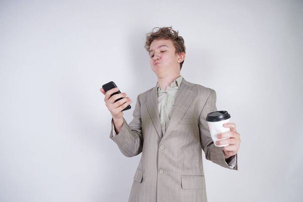 επιθετικός Καυκάσιος έφηβος φωνάζει στο έξυπνο τηλέφωνο και κρατά ένα χάρτινο φλιτζάνι καφέ στο χέρι του. φοιτητής στην άκρη σε λευκό φόντο στο Studio - Φωτογραφία, εικόνα