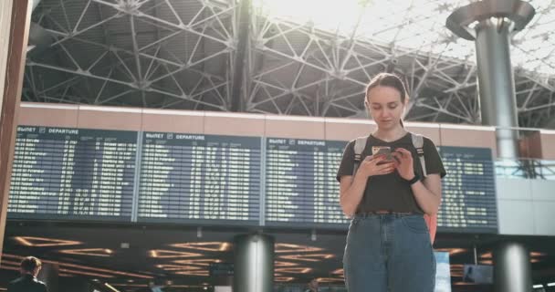 Eine junge Frau am Flughafen mit ihrem Smartphone in der Nähe der Anzeigetafel - Filmmaterial, Video
