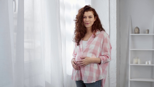 mooie zwangere jonge vrouw die de buik aanraakt, naar de camera kijkt en glimlacht thuis - Video