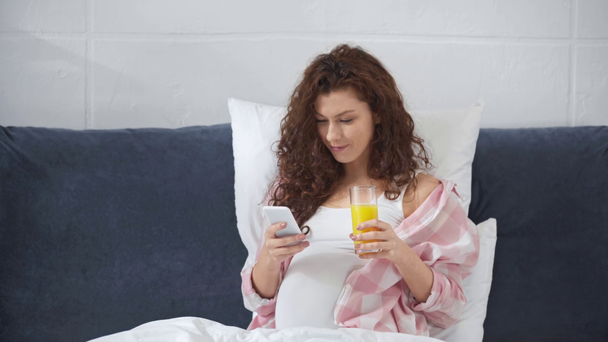 έγκυος νεαρή γυναίκα που βρίσκεται στο κρεβάτι, χρησιμοποιώντας smartphone και πίνοντας χυμό πορτοκάλι στο σπίτι - Πλάνα, βίντεο