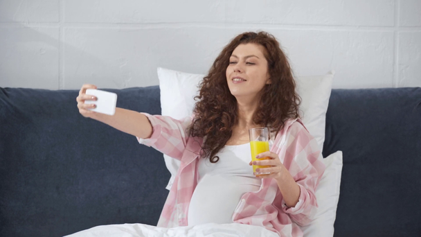 妊娠中の若い女性がベッドに横たわり、オレンジジュースを飲み、自宅でスマートフォンで自分撮りをする - 映像、動画