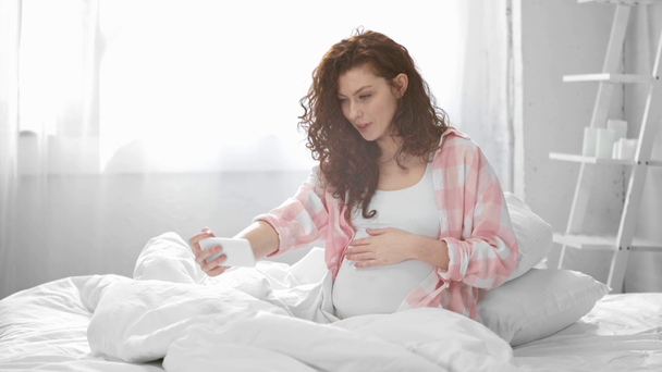 Schwangere junge Frau führt Videochat auf Smartphone, winkt und lächelt im Bett - Filmmaterial, Video