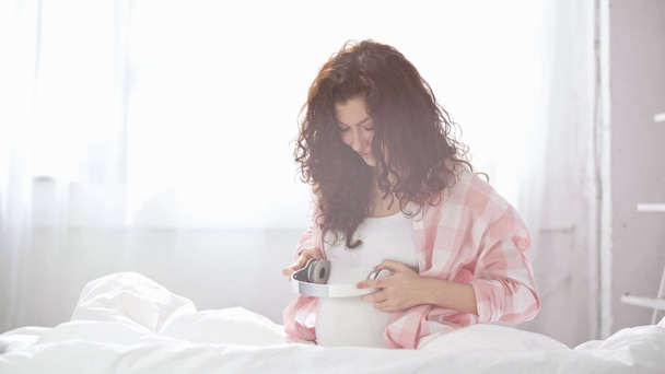 mooie zwangere jonge vrouw luisteren muziek, glimlachend en het houden van koptelefoon in de buurt van de buik in bed  - Video