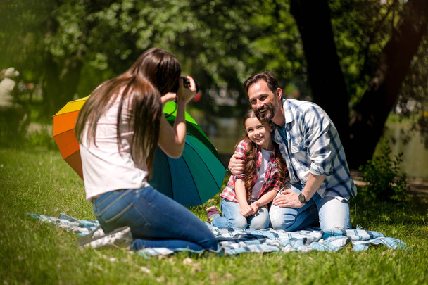 Молодая мать фотографирует мужа и дочь в парке. Отец обнимает свою дочь, сидя на одеяле. Красочный зонтик рядом с ними
. - Фото, изображение