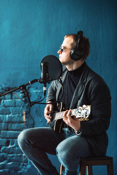 Nahaufnahme eines männlichen Sängers, der mit Kopfhörern auf einem Hocker sitzt und eine Gitarre spielt, die einen Song in einem Heimstudio aufnimmt - Foto, Bild