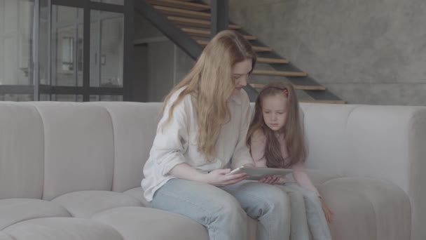 Beautiful young mother and her cute little daughter are using a tablet and smiling, sitting on sofa in the big house. Family relationships. - Кадри, відео