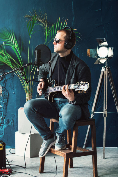 Крупный план певца, сидящего на стуле в наушниках с гитарой, записывающего трек в домашней студии
 - Фото, изображение