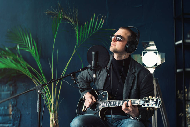 ホームスタジオでトラックを録音したギターを持つヘッドフォンで腰掛けに座っている男性歌手のクローズアップ - 写真・画像