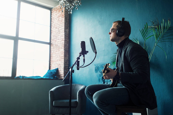 Close up de um cantor sentado em um banquinho em um fone de ouvido com uma guitarra gravando uma faixa em um estúdio em casa
 - Foto, Imagem