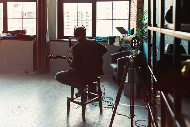 Άνθρωπος τραγουδίστρια κάθεται σε ένα σκαμνί σε ένα ακουστικό με μια κιθάρα που καταγράφει ένα κομμάτι σε ένα στούντιο σπίτι - Φωτογραφία, εικόνα