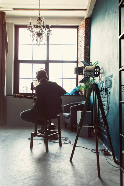 Hombre cantante sentado en un taburete en unos auriculares con una guitarra grabando una pista en un estudio en casa
 - Foto, imagen