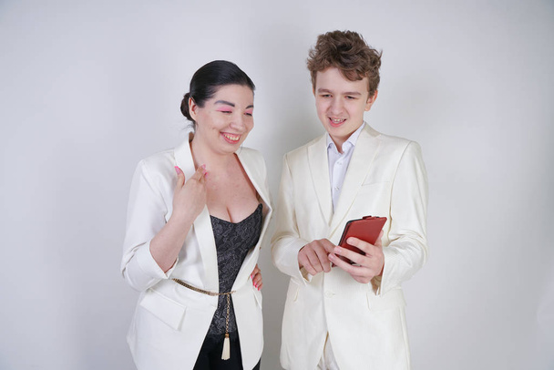caucasian teenager in a white business suit stands next to friendly adult asian woman and holds a phone on a white background - Photo, Image