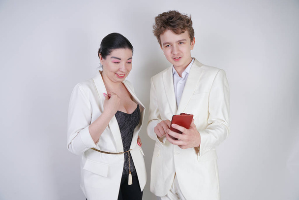 白いビジネススーツを着た白人のティーンエイジャーは、フレンドリーな大人のアジアの女性の隣に立って、白い背景に携帯電話を保持しています - 写真・画像