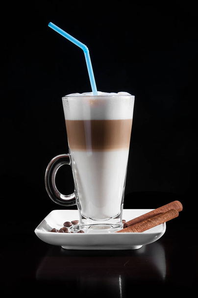 Latte macchiato in a glass with white plate, spoon, decorated with coffee beans, cinnamon. Black background. - Photo, Image