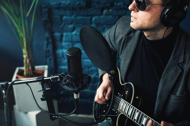 Крупный план певца, сидящего на стуле в наушниках с гитарой, записывающего трек в домашней студии
 - Фото, изображение