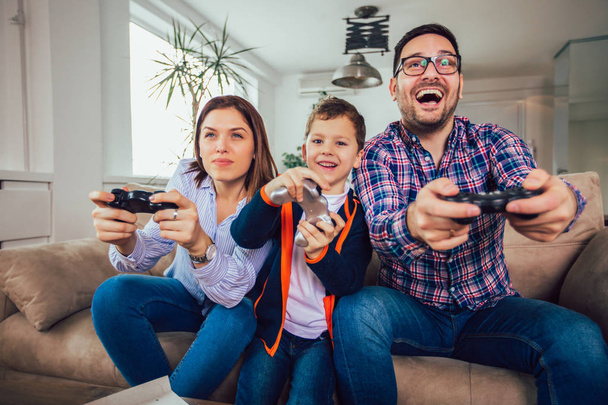 Счастливая семья, сидящая на диване, играющая в видеоигры и кушающая
 - Фото, изображение