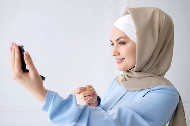 Μουσουλμανική γυναίκα εφαρμόζει συμπαγή σκόνη κοιτάζοντας καθρέφτη. - Φωτογραφία, εικόνα